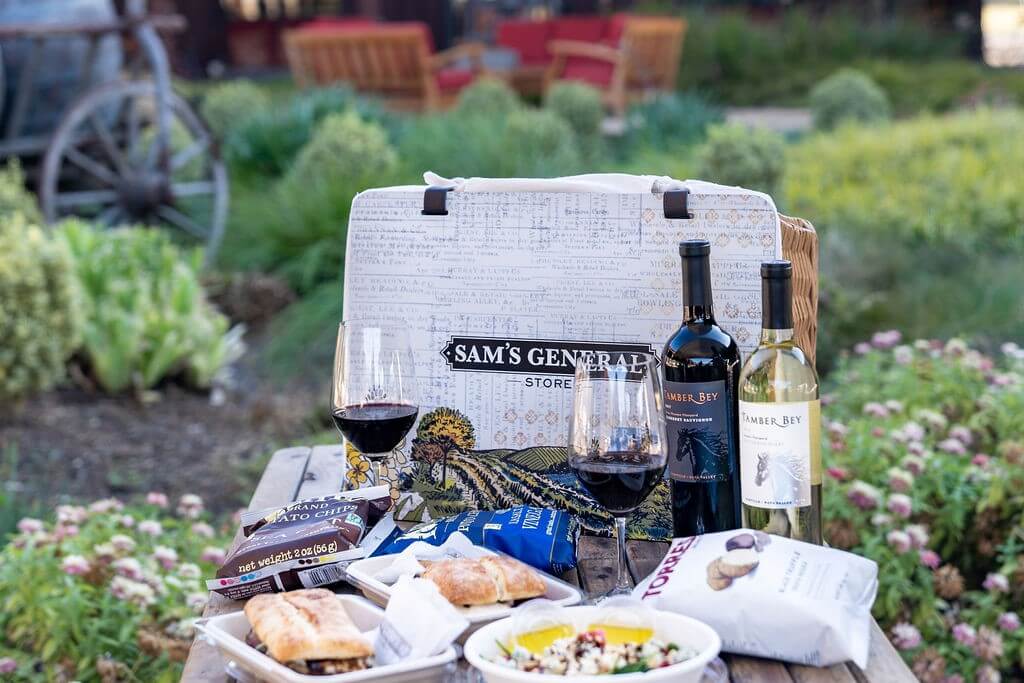 An open picnic basket brimming with food and wine sits in a sunny garden.