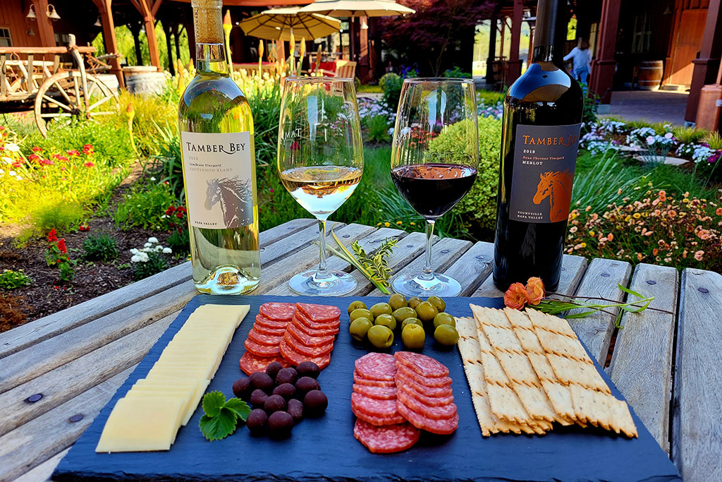 Salami and crackers on a blue slatewith a glass and bottle of red and white wine on a table with flowers and umbrellas in the background