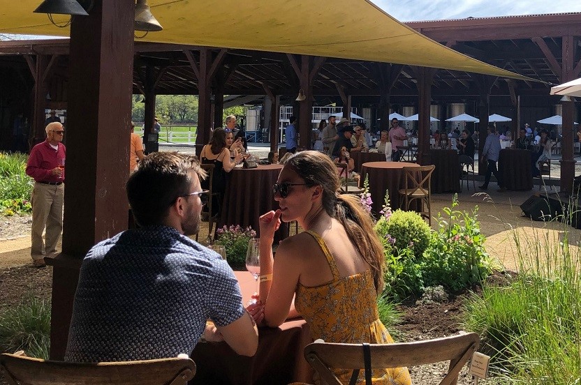 Couple sitting outdoors under an umbrella enjoying wine with additional people standing in the background.